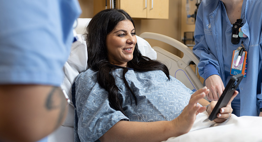A young pregnant woman with brown hair rests in a hospital bed. An RN stands on either side of her, faces not visible. One is helping the pregnant woman access her medical info on a tablet. 