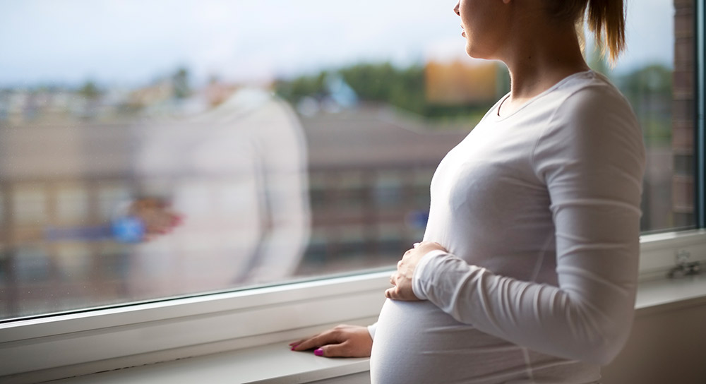 Una madre joven toca su panza y mira a través de una ventana a la distancia.