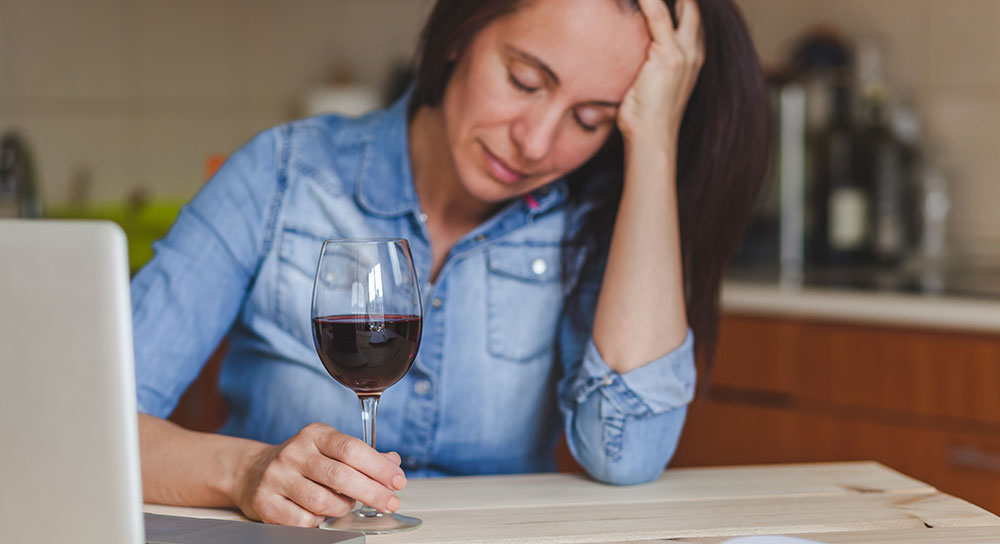 A white woman with brown hair holds the stem of a glass filled with red wine. Her head is in her hands and she is distraught.