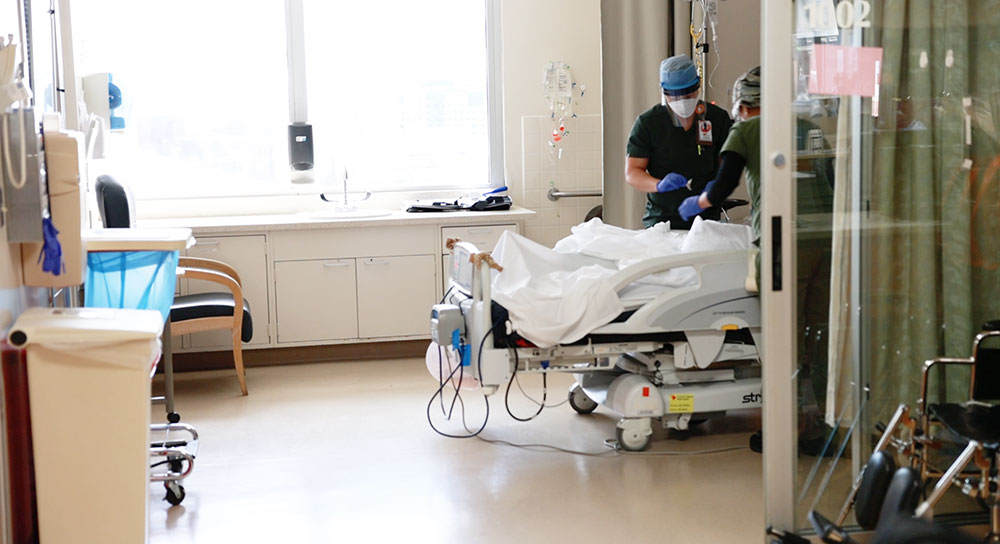 Plano amplio de una habitación de hospital con dos profesionales de la salud con máscaras atendiendo a un paciente cuyo rostro no es visible.