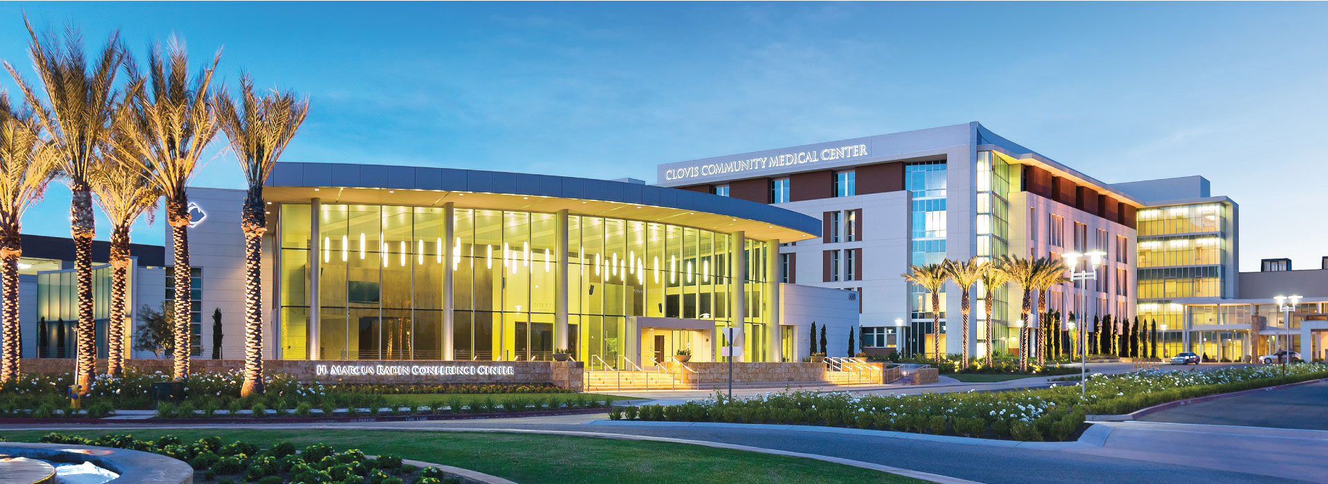 Exterior shot of Clovis Community Medical Center at dusk. The background sky is dark blue and the yellow lights in the facility glow brightly.