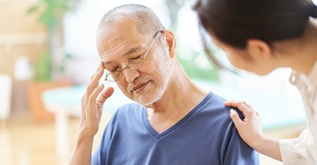 An older Asian man is holding his head in confusion while a younger woman places a concerned hand on his shoulder.