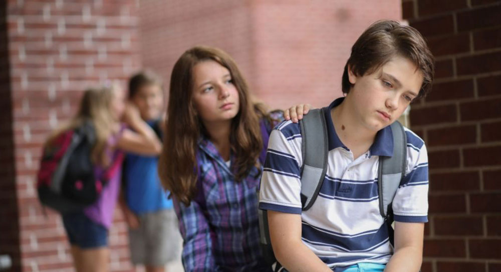 Two white kids, a boy and a girl, high school-aged, are at school. The girl places a supportive hand on the boy's shoulder. 