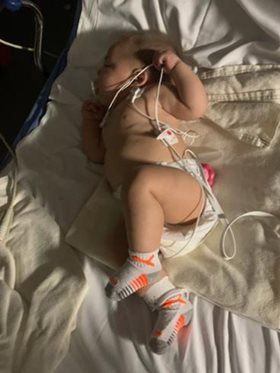 6-month-old Kruz Rojas lies in a hospital bed with tubes connected to his chest and mouth