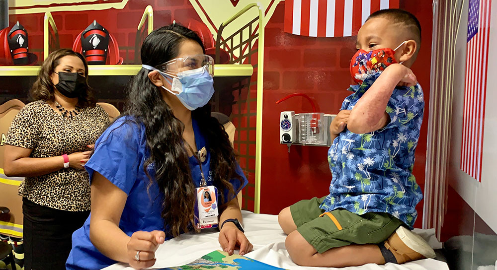 A young boy sits on an exam table facing his mom and a child life specialist. All are wearing face masks.
