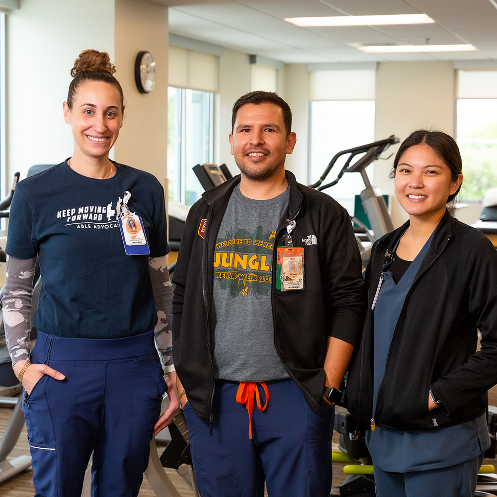 Tres terapeutas físicos están en un gimnasio: una mujer blanca, un hombre blanco y una mujer asiática.