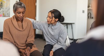 a young Black woman consoles an older Black woman who is sitting in a chair clasping her hands together