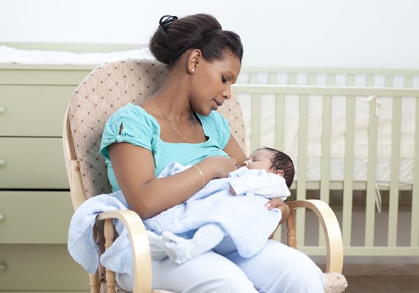 A woman, young and Black, breastfeeds her newborn child in a baby's nursery