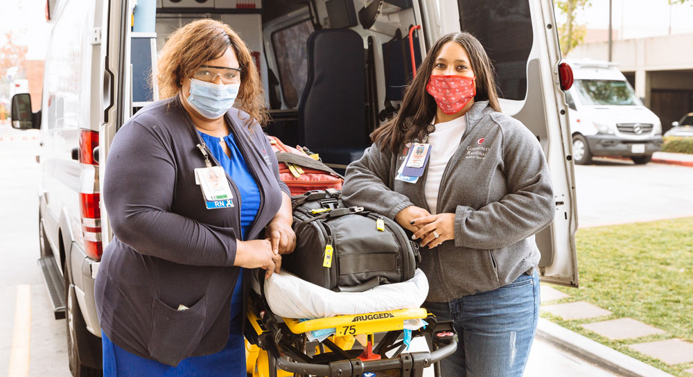 Two masked nurses, both female and masked, stand in front of an ambulance and gurney