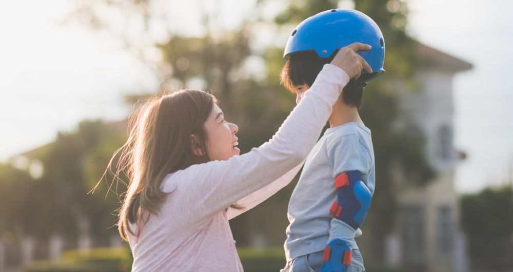 A mother and her child, both Asian, smile as mom places a helmet on her son's head.