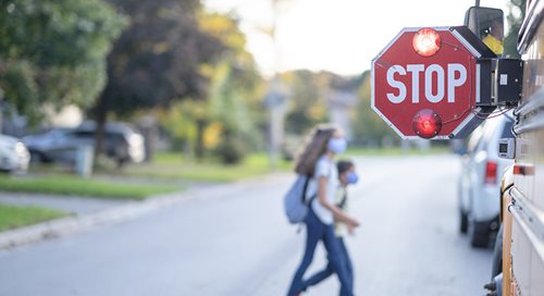red stop sign attached to school bus in foreground. In the background, two children -- one a girl, one a boy, both wearing masks -- cross the street