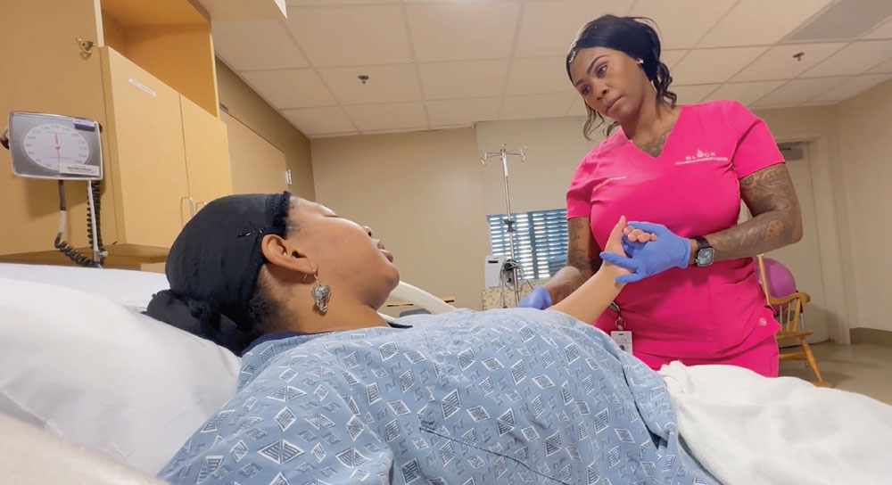 A doula in training holds the hand of a pregnant patient who is laying in bed