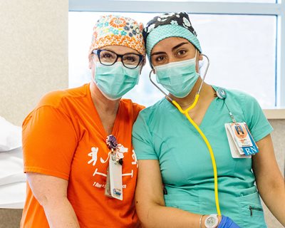 two nurses in scrub caps and gloves stand side by side and smile