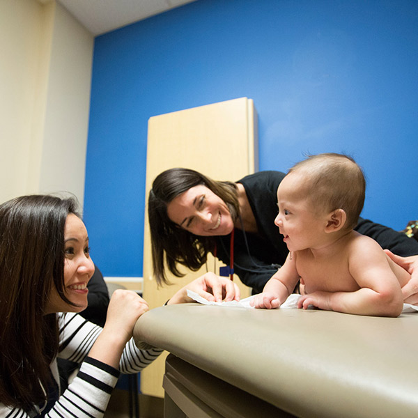 Una madre joven le sonríe a su bebé, que está siendo sostenido por una trabajadora de la salud en una sala de examen.