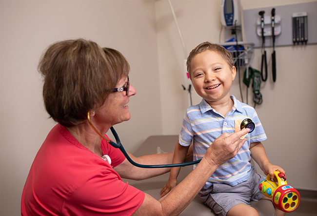 A female nurse in red holds a stethoscope to a young boy who is smiling and holding a toy.