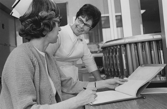 A black-and-white photo of two nurses chatting over a chart.
